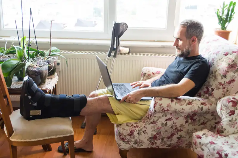 Man wearing walking boot for achilles tendon treatment is using laptop at home. Injured man with crutches.