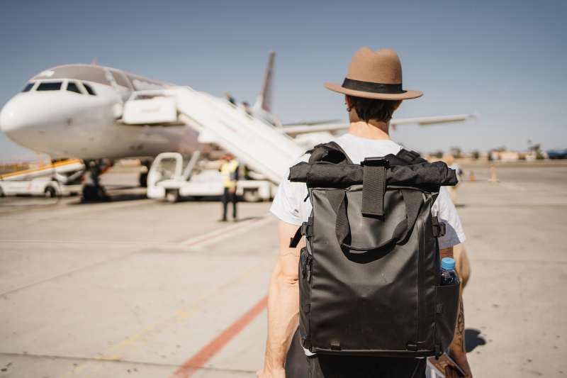 Young white man with hat and suitcase walking through the airport on the runway towards his airplane
