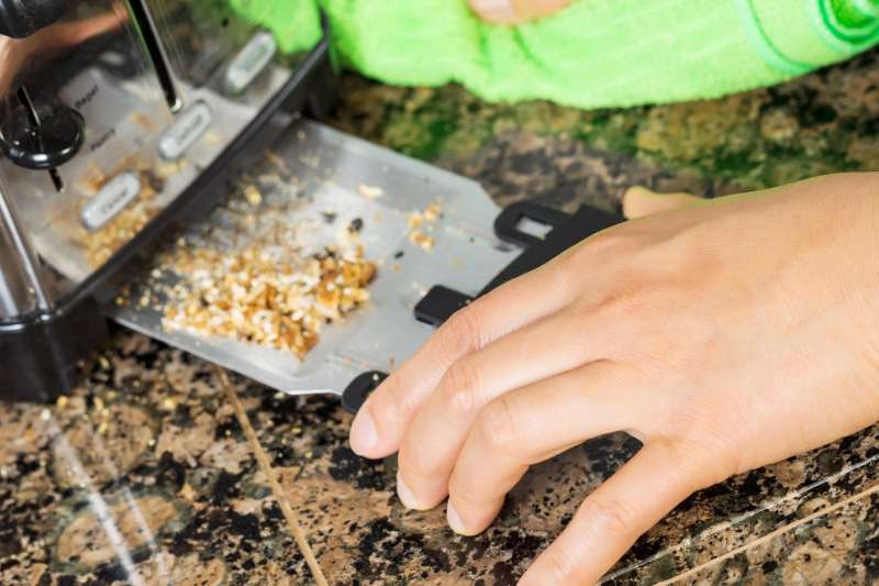 Photo of female hand cleaning bottom of kitchen toaster tray with green microfiber rag with stone counter top in background