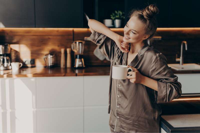 Young pretty relaxed woman in comfy pajama stretching from sleep early in morning while holding cup of coffee in her hand, standing in stylish kitchen interior.