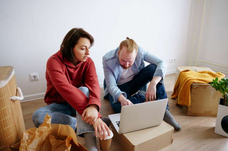 Couple sitting on the floor of a living room with moving boxes, looking at a laptop