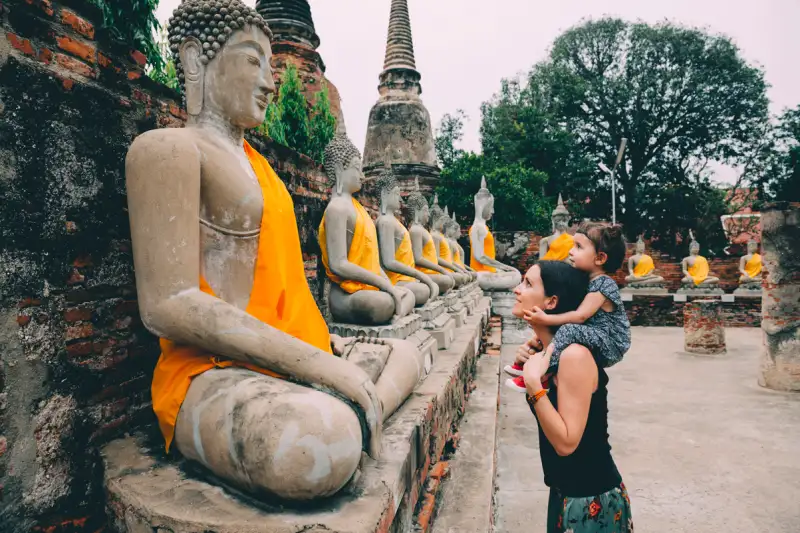 Young Woman, carrying her daughter on her shoulders, looking at a Buddha statue in Thailand