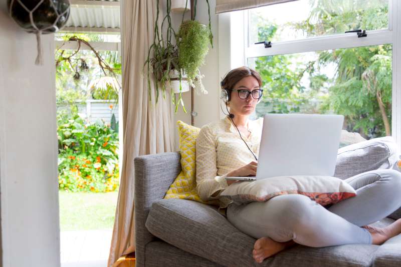 Young woman working from home, using her laptop sitting on a couch next to a window