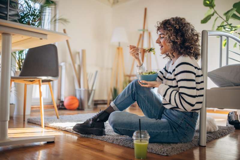 Young woman sitting on the floor at home and eating a salad and smoothie.