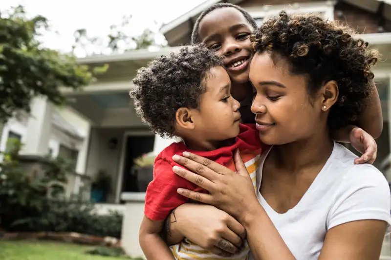 Mother embracing her two young children in-front of a suburban house