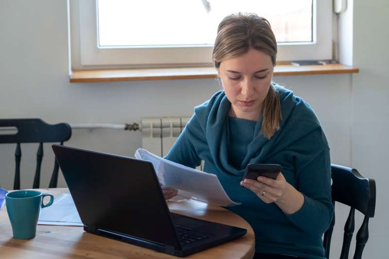 Woman holding document while looking at her phone near a laptop.