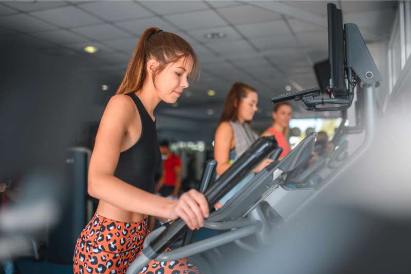 Side view personal perspective of three young female gym enthusiasts working out on elliptical trainers.