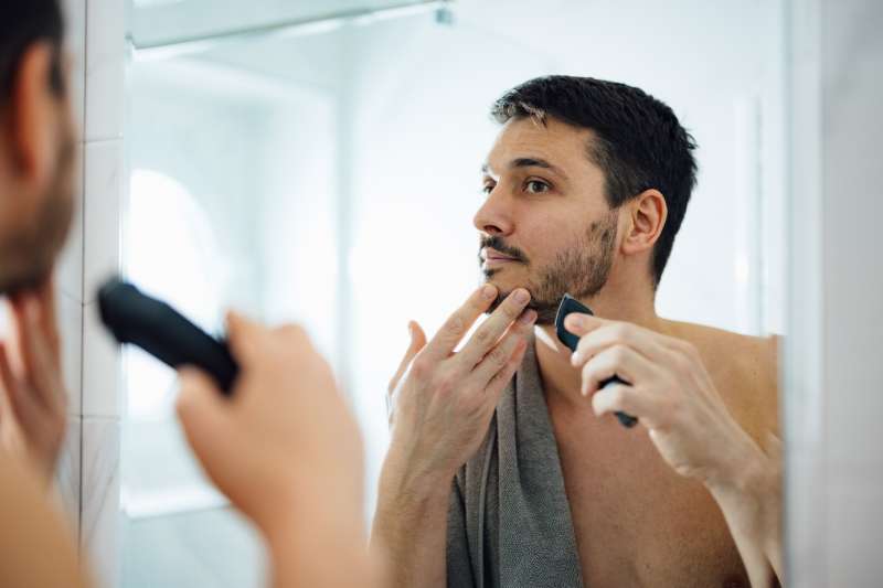Close up photo of serious man trimming his beard with a trimmer in front of a bathroom mirror in the morning.