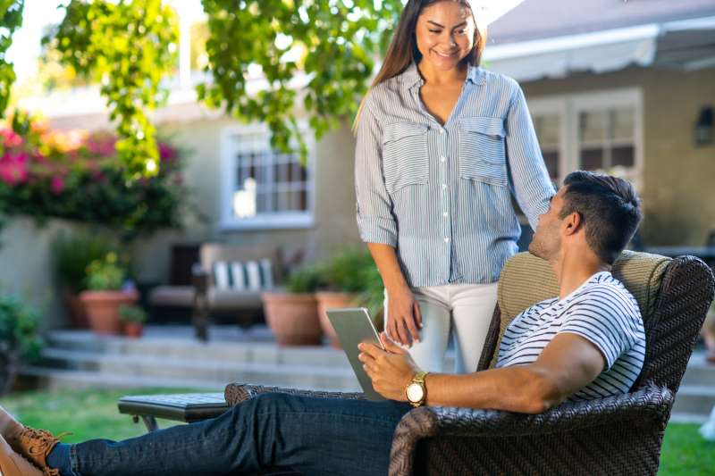 A young nicely dressed hispanic couple relax in their suburban backyard while the man shops on his tablet.