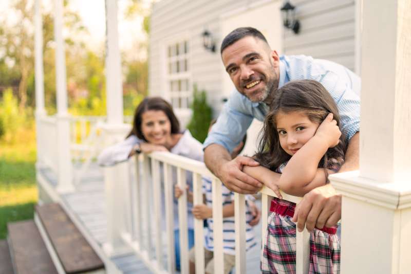 A family of four standing on the front porch and leaning on the railings while smiling at the camera outside on a bright day