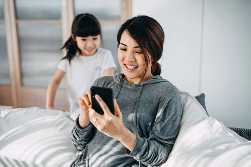 Busy young Asian mother sitting in bed using smartphone while little daughter is distracting her and seeking for attention