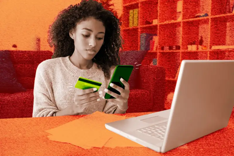 Woman holding a smartphone in one hand and a credit card in the other, sitting in-front of her computer