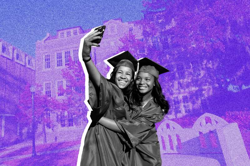 Two students, wearing a cap and gown, taking a selfie in-front of a college building